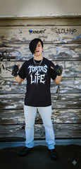 Person wearing a black t-shirt with 'Tortas4Life' text, standing against a rustic wooden wall with graffiti.